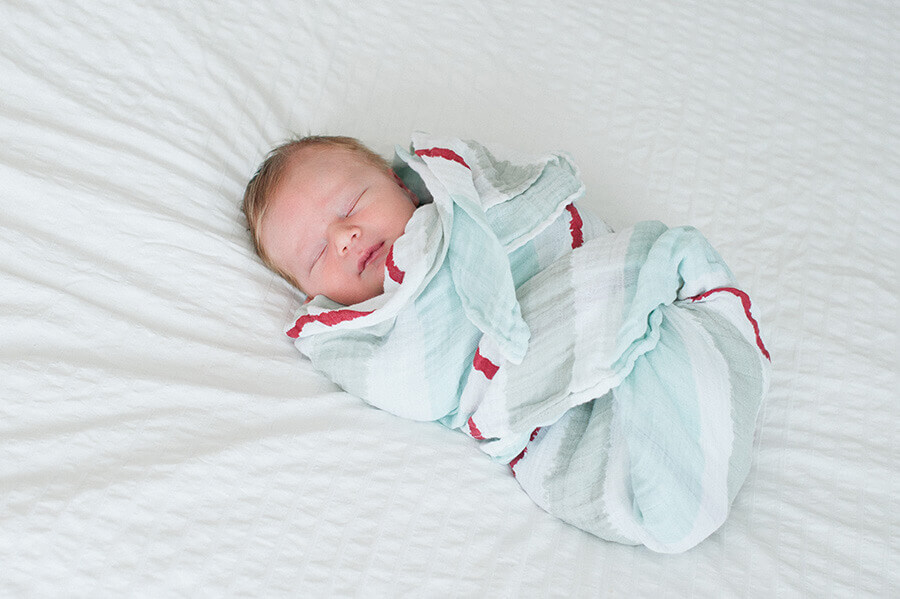 A newborn baby bundled in a blue blanket with red trim sleeps on a soft bed dreaming of the new world around him. Photo by AMBphoto
